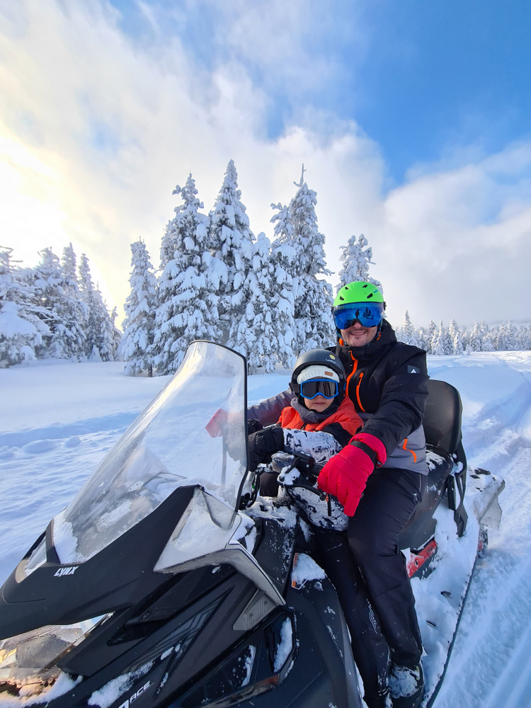 Adult și copil pe un snowmobil, lângă brazi înzăpeziți.
