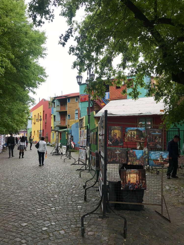 Stradă pietonală umbrită de arbori, cu tarabe și clădiri colorate.