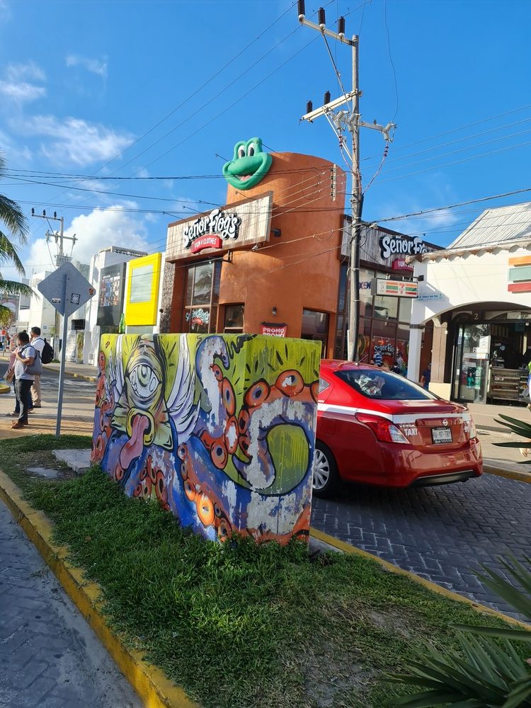 Colț de stradă cu panou pictat, mașină și clădiri colorate.