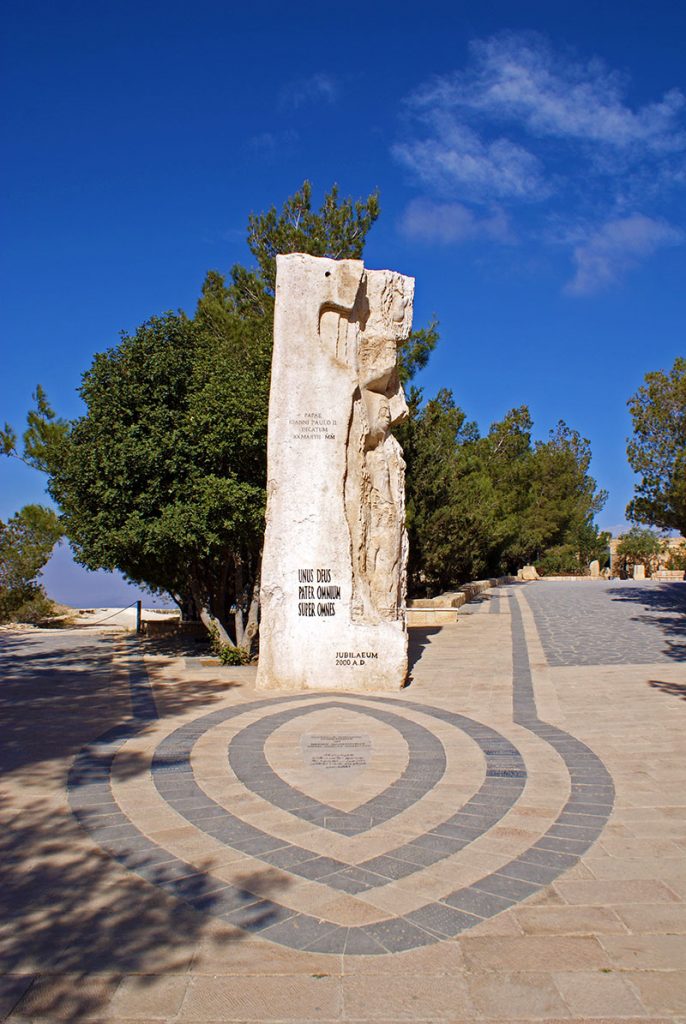 Monument vertical din piatră, într-o piațetă cu pavaj circular.