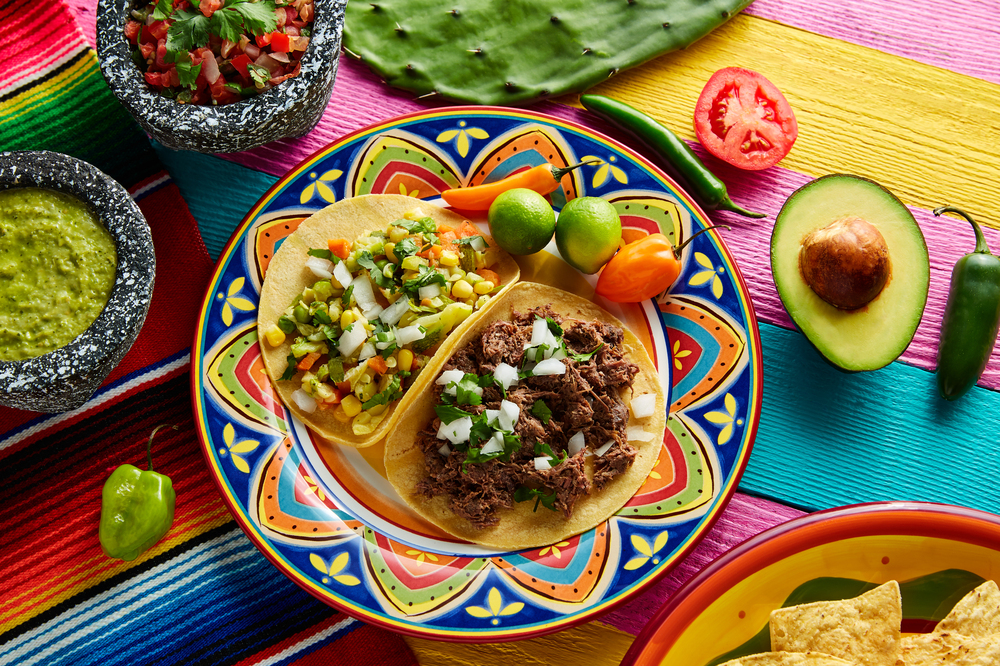 Tacos cu garnituri, avocado și sosuri pe o masă colorată.