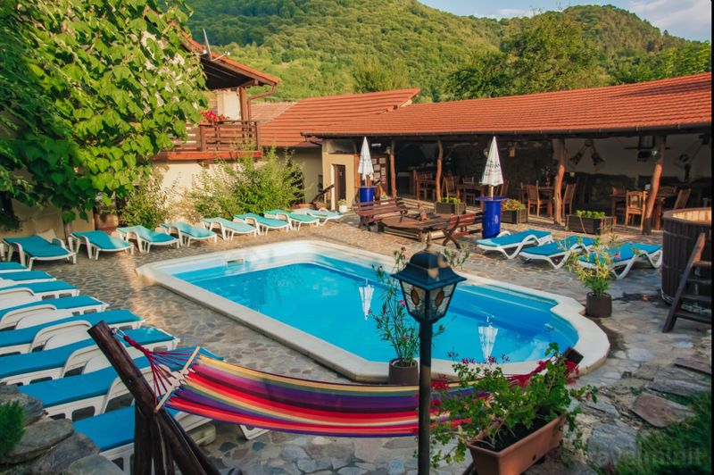 Piscină exterioară cu șezlonguri, lângă clădiri și dealuri împădurite.