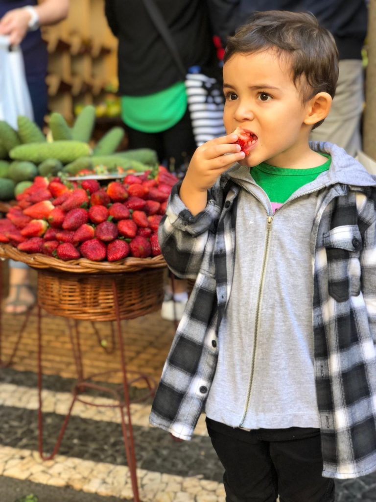 Copil gustând un fruct lângă o tarabă.