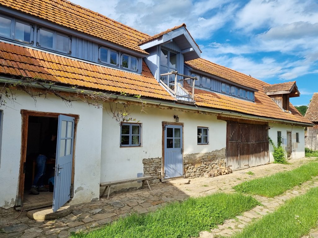 Clădire rurală albă cu acoperiș roșcat și uși albastre.