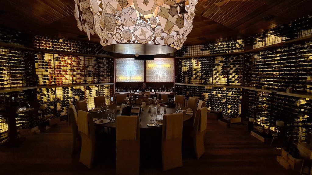 Masă lungă sub candelabru într-o încăpere cu rafturi de vin.