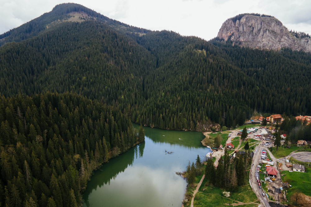 Lac și clădiri între munți împăduriți, văzute de sus.
