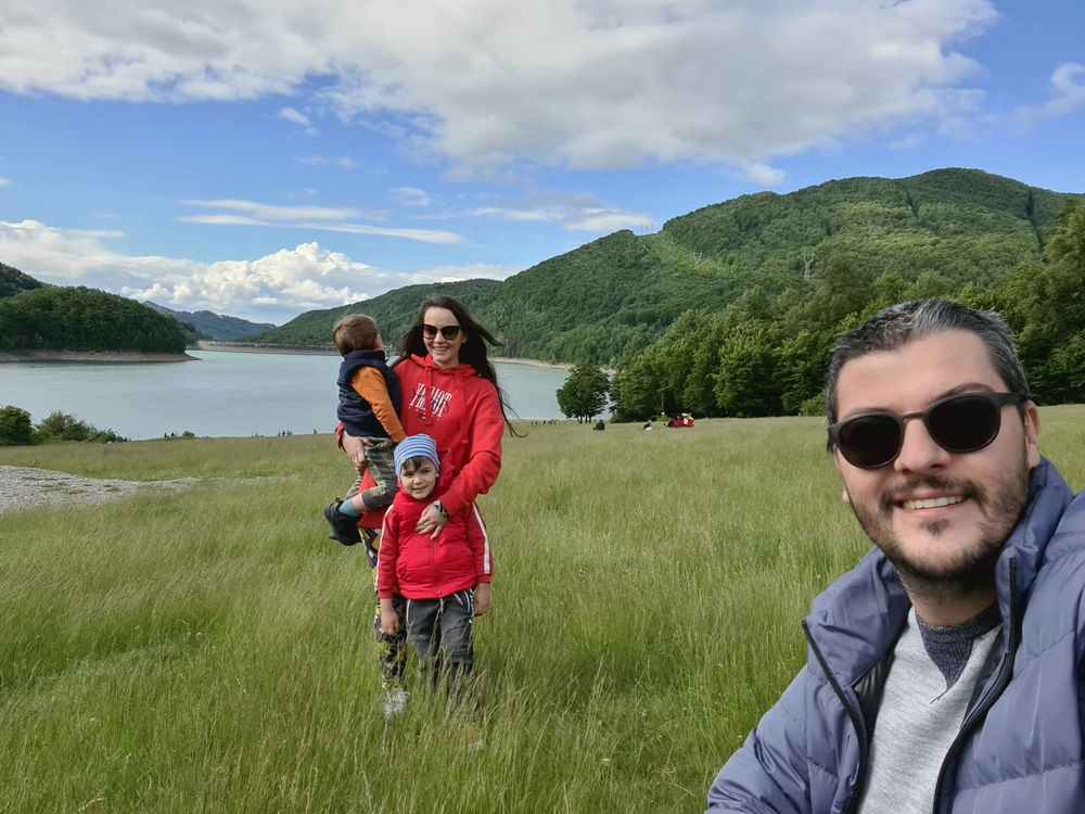 Adulți și copii lângă un lac, cu dealuri împădurite în fundal.