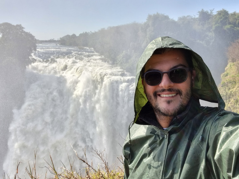 Bărbat cu ochelari și pelerină, în fața unei cascade.