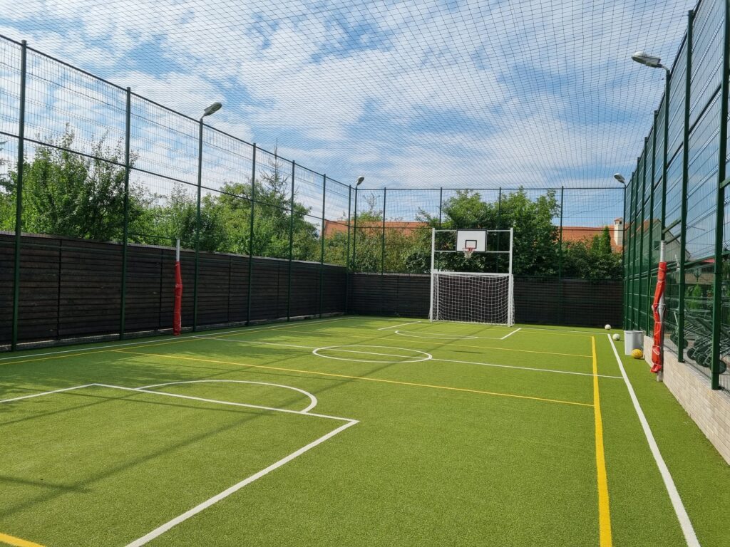 Teren sportiv verde, împrejmuit cu plasă, cu poartă și coș.
