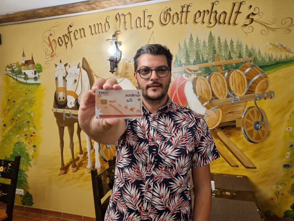 Bărbat cu ochelari ridicând o halbă, în fața unui perete pictat.