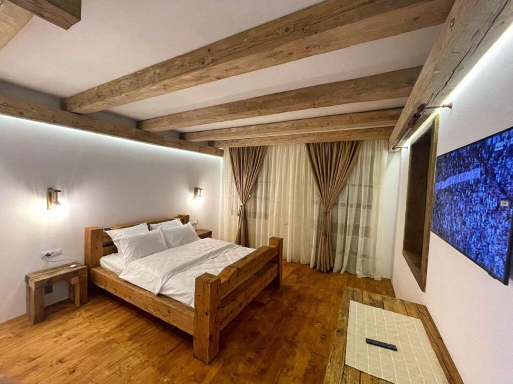 Dormitor cu pat din lemn, grinzi aparente și televizor mare.