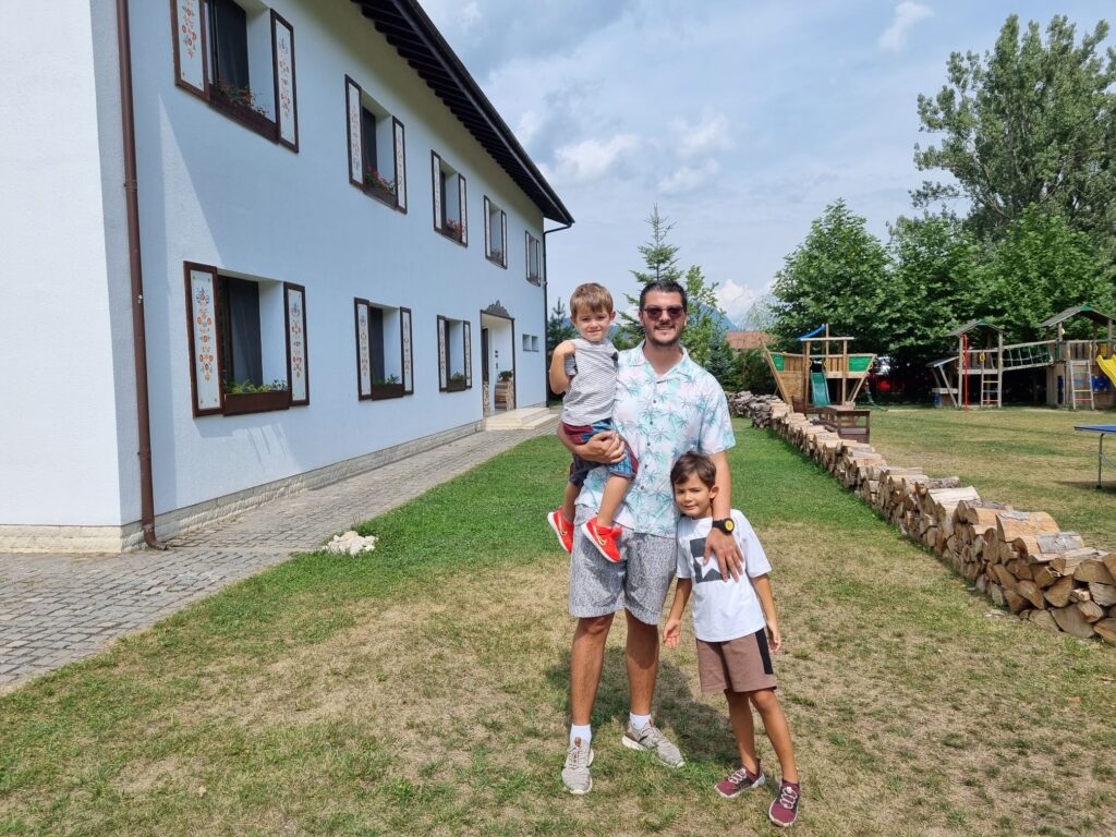 Bărbat și doi copii pe gazon, lângă o clădire albă.