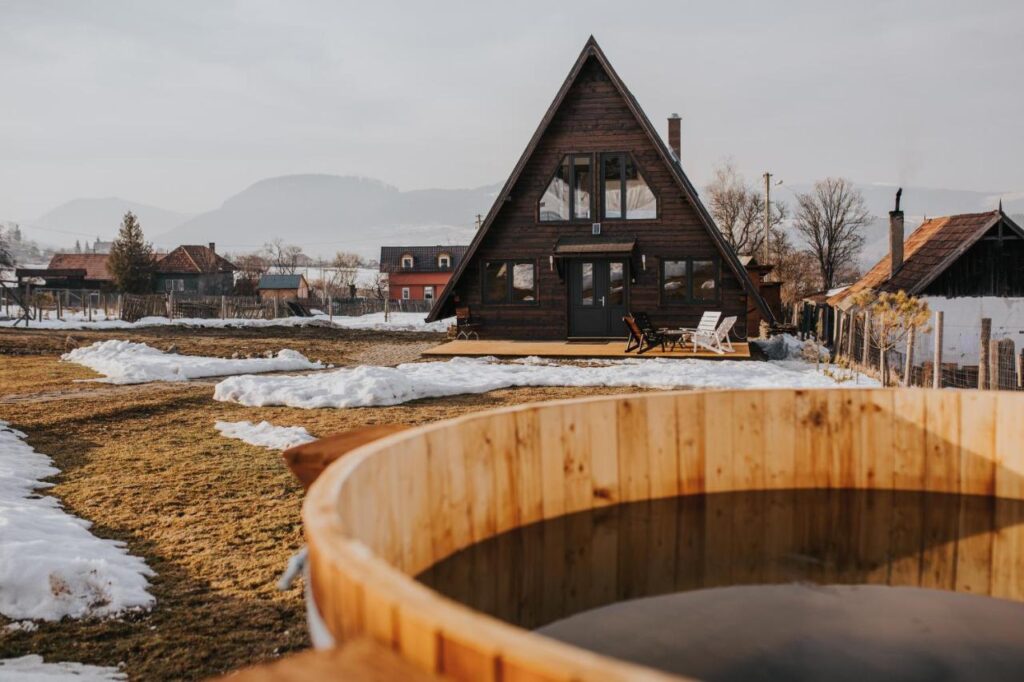 Ciubăr din lemn în fața unei cabane triunghiulare, între petice de zăpadă.