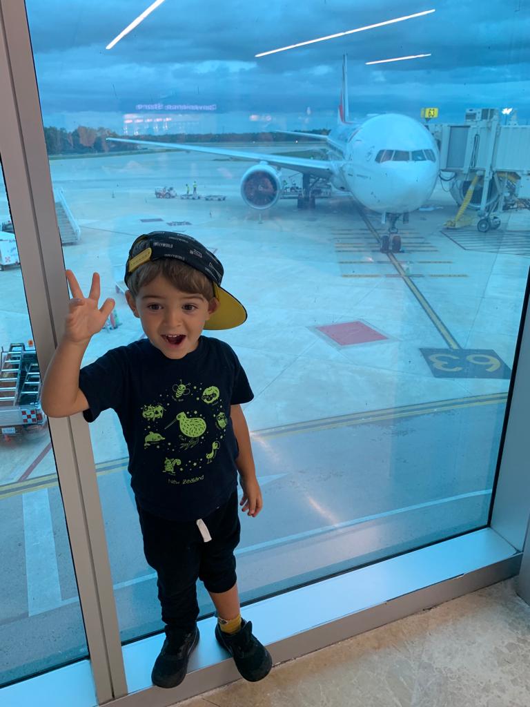 Copil lângă o fereastră de aeroport, cu un avion în fundal.