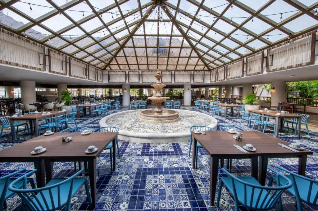 Restaurant cu acoperiș vitrat înalt, fântână centrală și mese aranjate.