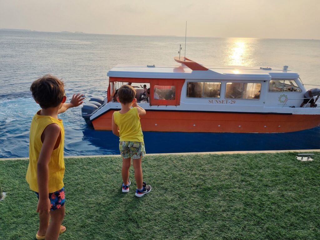 Doi copii pe malul apei, în fața unei ambarcațiuni.