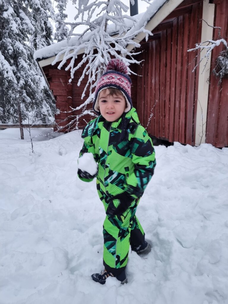Copil în combinezon verde, pe zăpadă, lângă o clădire din lemn.