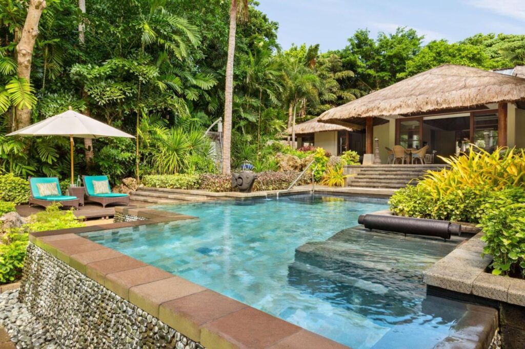 Piscină lângă o vilă cu acoperiș de paie și vegetație tropicală.