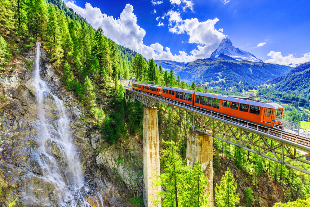 Tren roșu traversând un viaduct, lângă o cascadă și munți.
