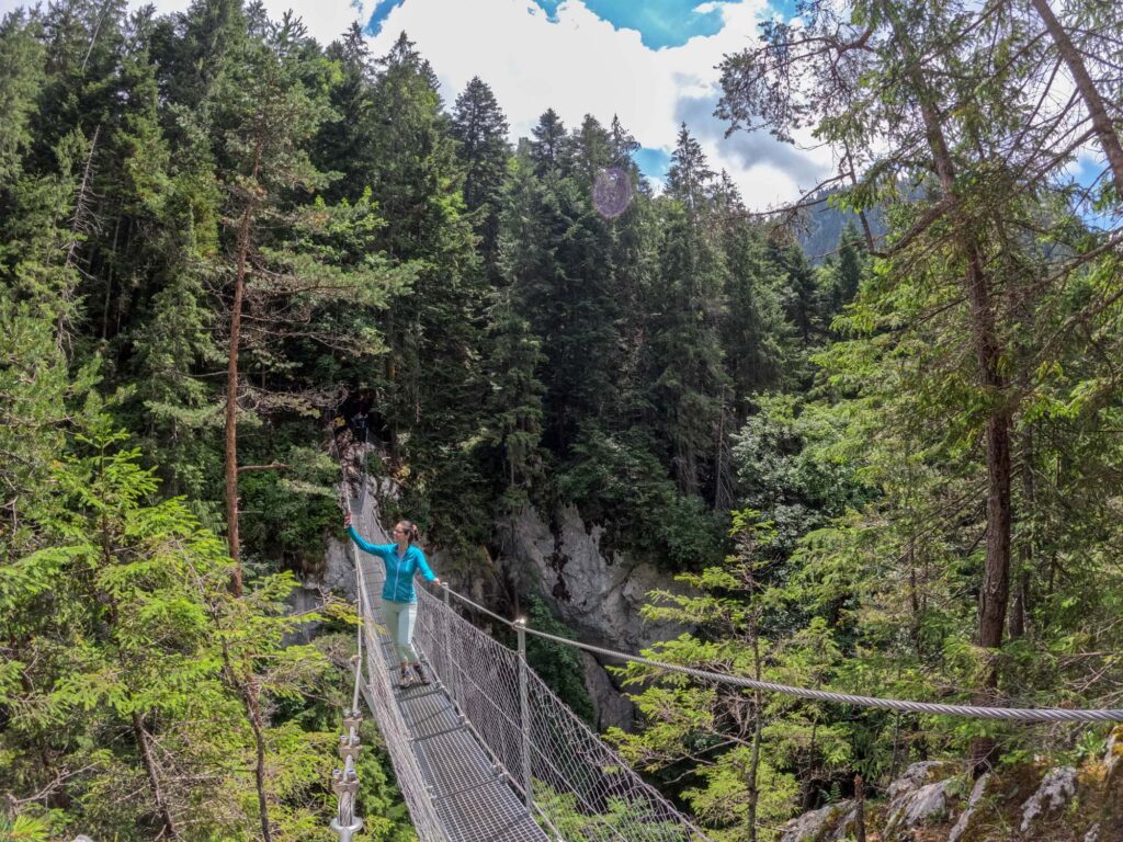 Persoană traversând o punte suspendată peste o vale împădurită.