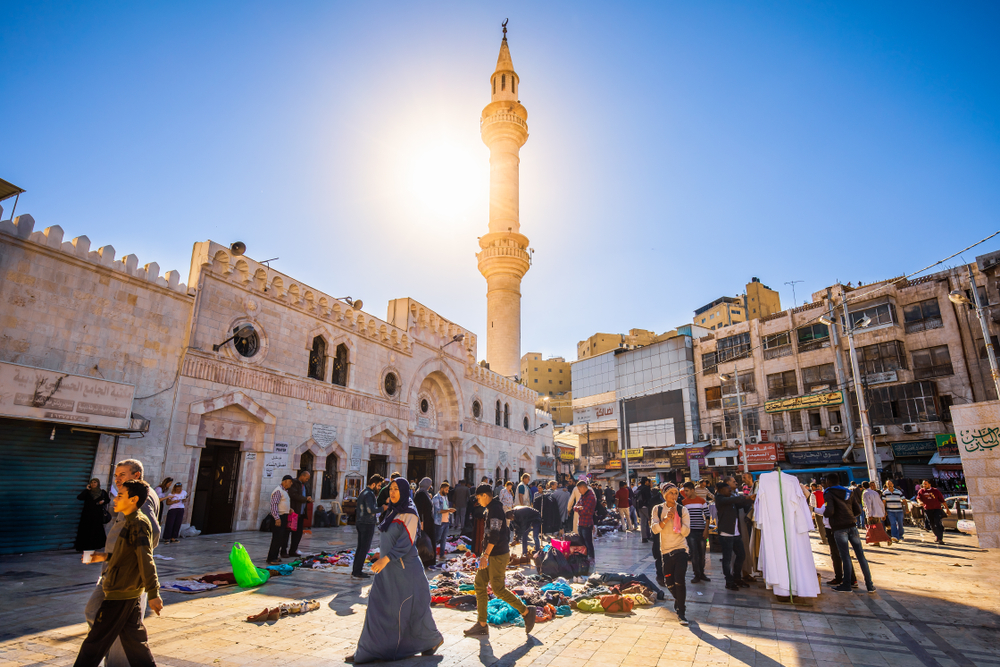 Pietoni și tarabe lângă o moschee cu minaret luminat de soare.