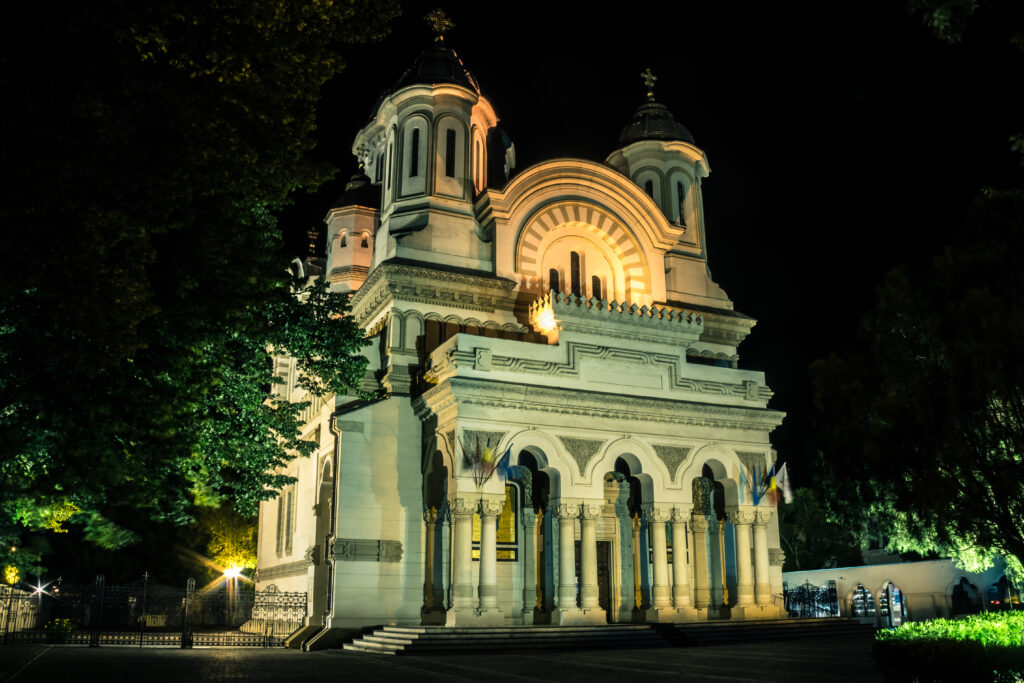 Biserică iluminată noaptea, cu arcade și turle, între arbori.