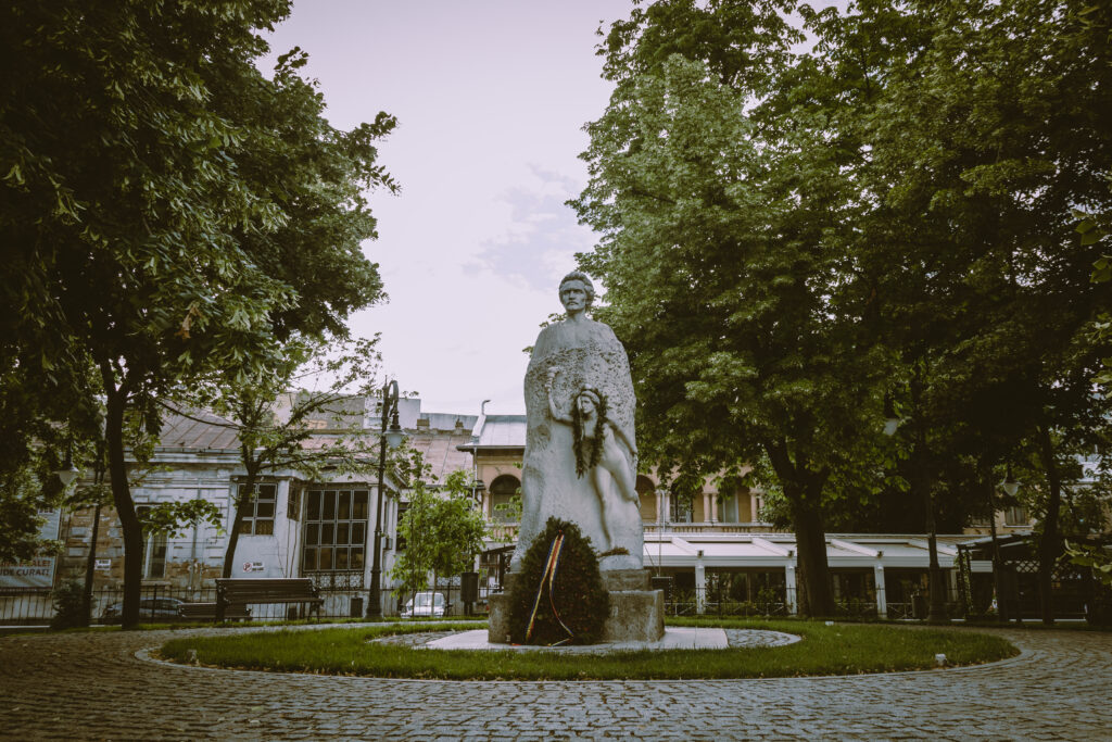 Statuie pe soclu într-un parc cu arbori și o clădire în fundal.