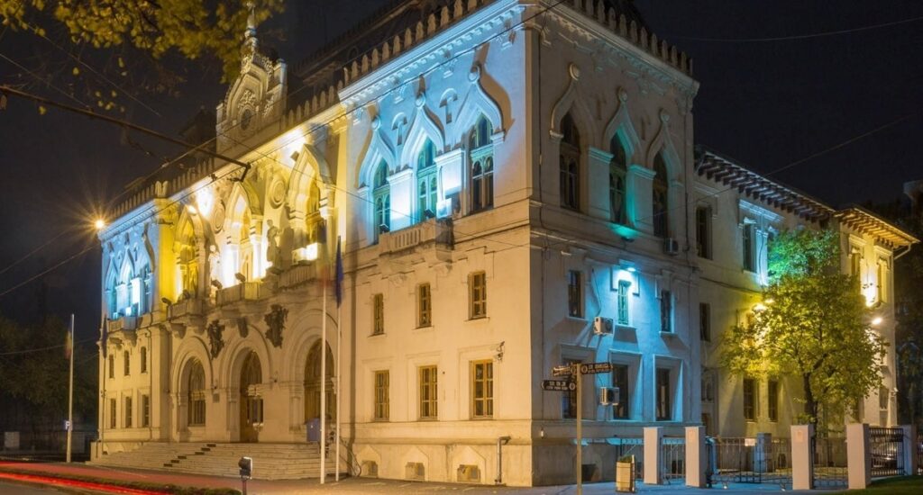 Clădire istorică iluminată noaptea în nuanțe de galben și albastru.