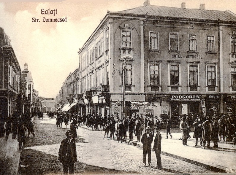 Fotografie veche cu pietoni pe o stradă mărginită de clădiri și prăvălii.