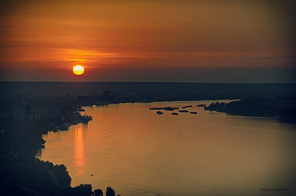 Soare portocaliu deasupra unui fluviu, cu reflexie luminoasă pe apă.