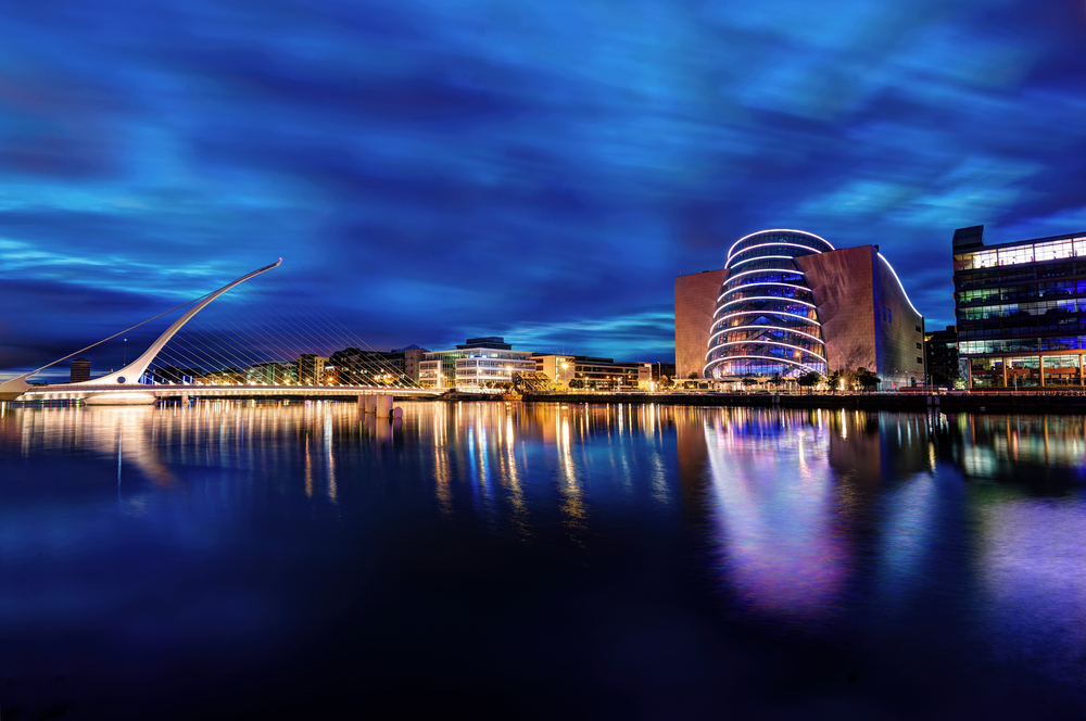 Pod cu cabluri și clădiri moderne iluminate, reflectate în apă noaptea.