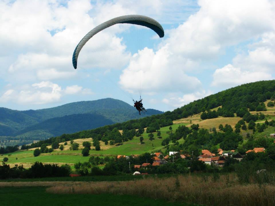 Parapantă în zbor deasupra unei văi cu pajiști și case.