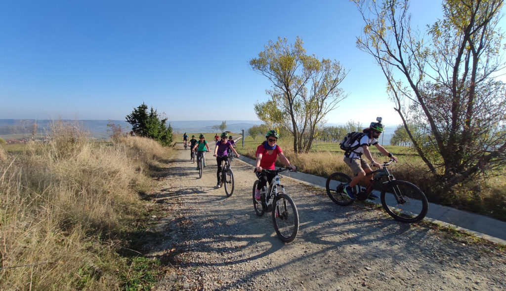 Grup de bicicliști pe un drum rural, între iarbă și arbori.