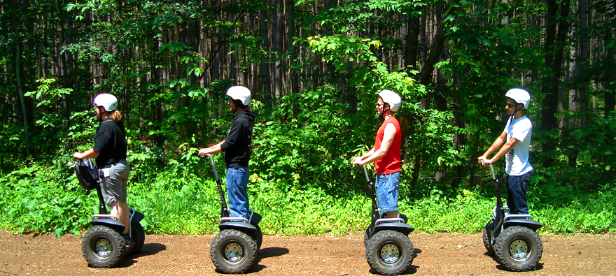 Patru persoane cu căști pe vehicule Segway, pe un drum prin pădure.