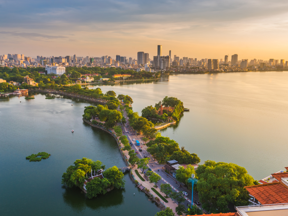 Lac urban cu insulițe verzi, alee pe mal și clădiri în fundal.