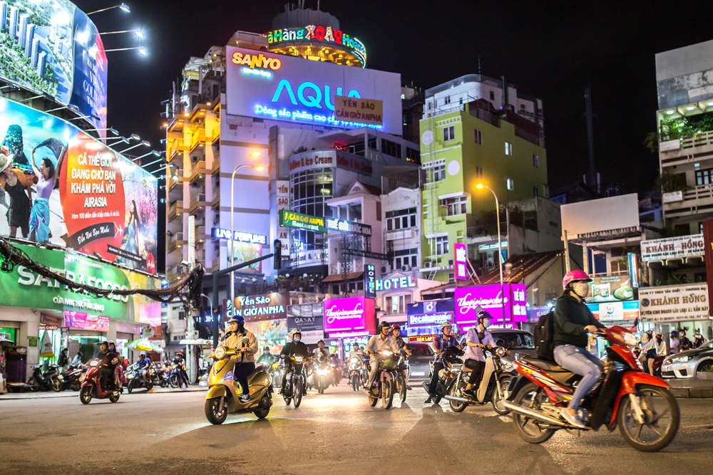 Intersecție aglomerată noaptea, cu motociclete și fațade pline de reclame luminoase.