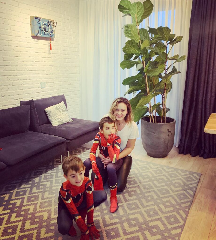 Femeie și doi copii într-un living cu canapea violet și plante mari.