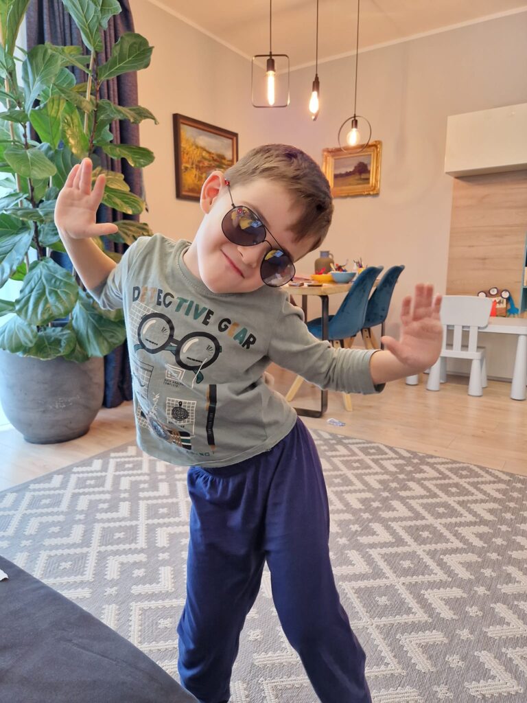 Copil cu ochelari de soare și brațele ridicate, într-o cameră.