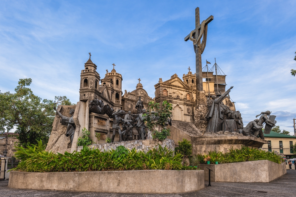 Monument cu cruce și sculpturi, înconjurat de vegetație.