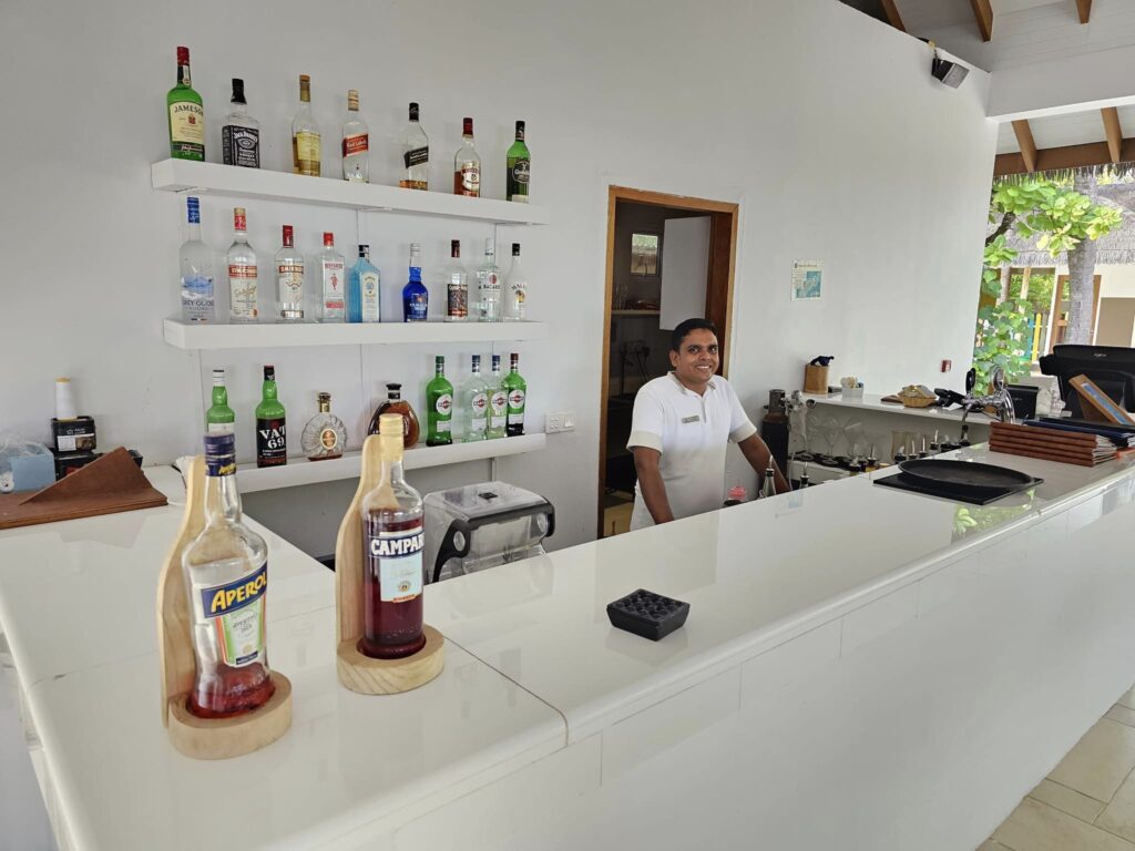 Barman în spatele unei tejghele albe, lângă rafturi cu sticle.