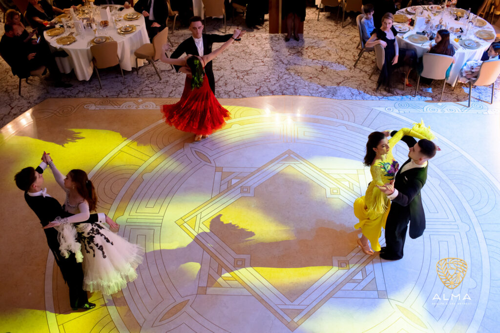 Perechi de dansatori în ținute elegante, pe un ring luminat.