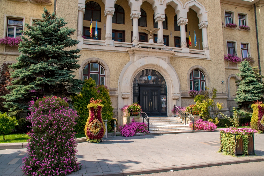 Intrare arcuită într-o clădire istorică, înconjurată de flori și conifere.