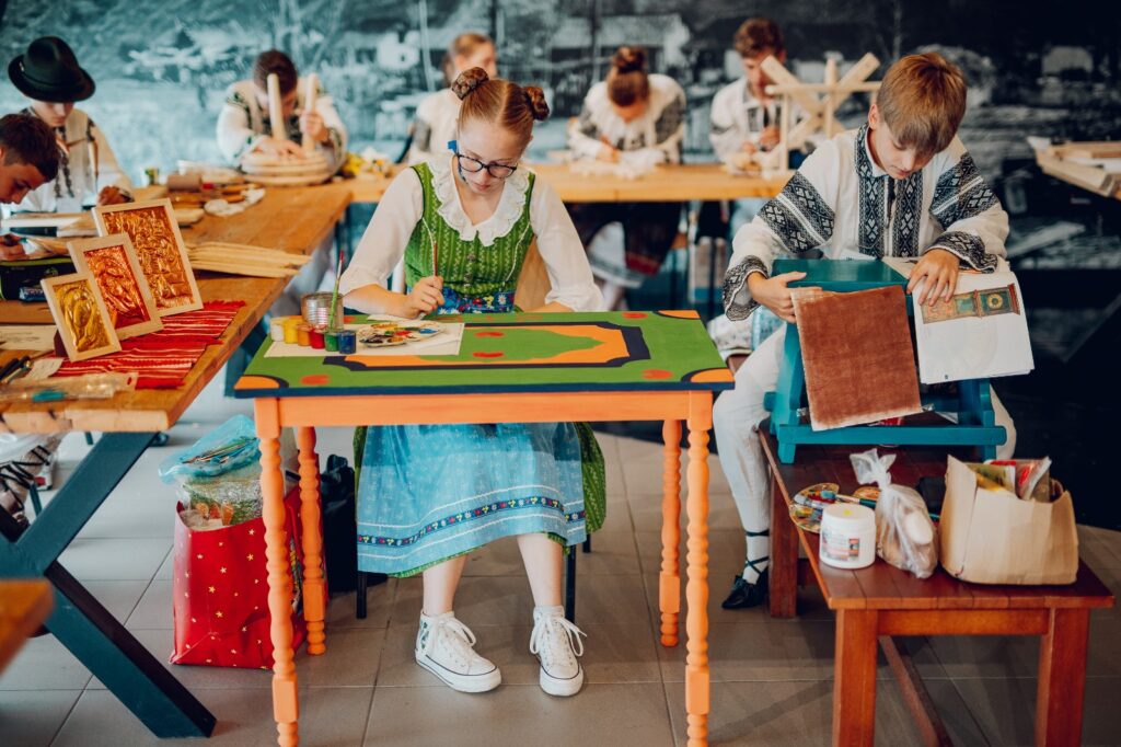 Participanți așezați la mese pictate, într-un atelier în aer liber