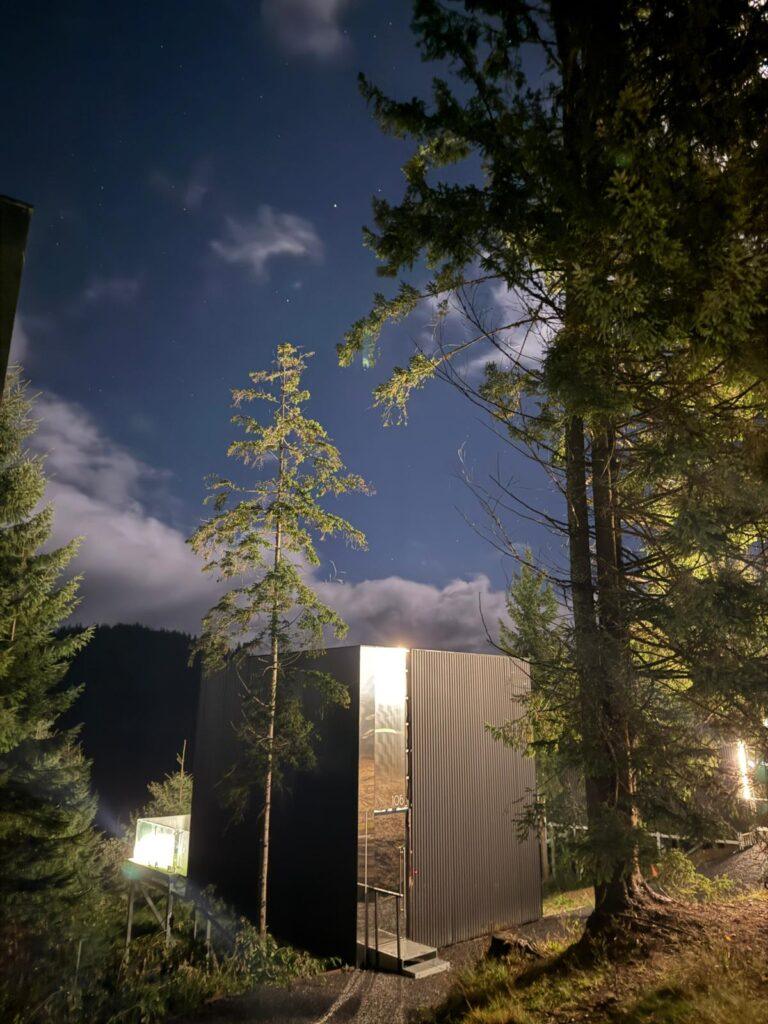 Cabană cu fațadă vitrată între arbori, la lăsarea serii
