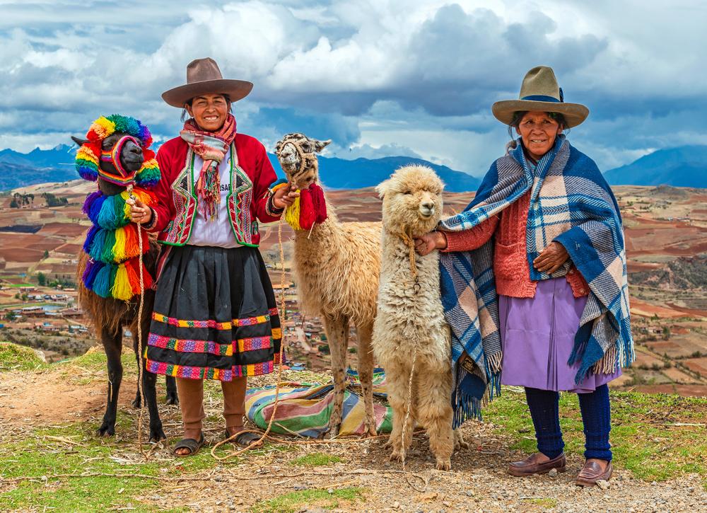 Două femei în haine tradiționale lângă alpaca