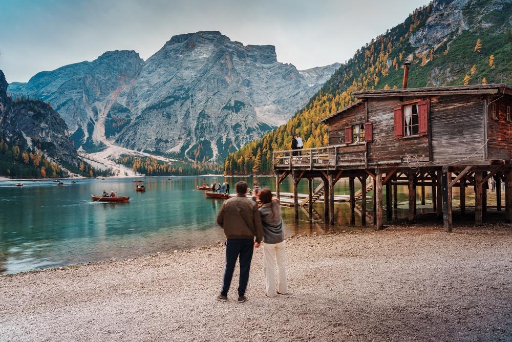 Persoană pe malul unui lac, lângă cabane și munți
